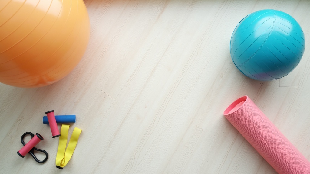 A flat lay of exercise equipment including a stability ball, yoga mat, and medicine ball on a wooden floor in natural light